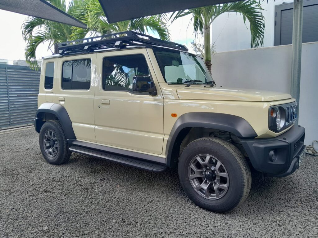 Suzuki Jimny 5-door 4×4 for rent in Mauritius from Liberta Car Rental—bright yellow extended-wheelbase Jimny parked on a sandy cliff road overlooking the Indian Ocean, surfboards strapped to the roof rack