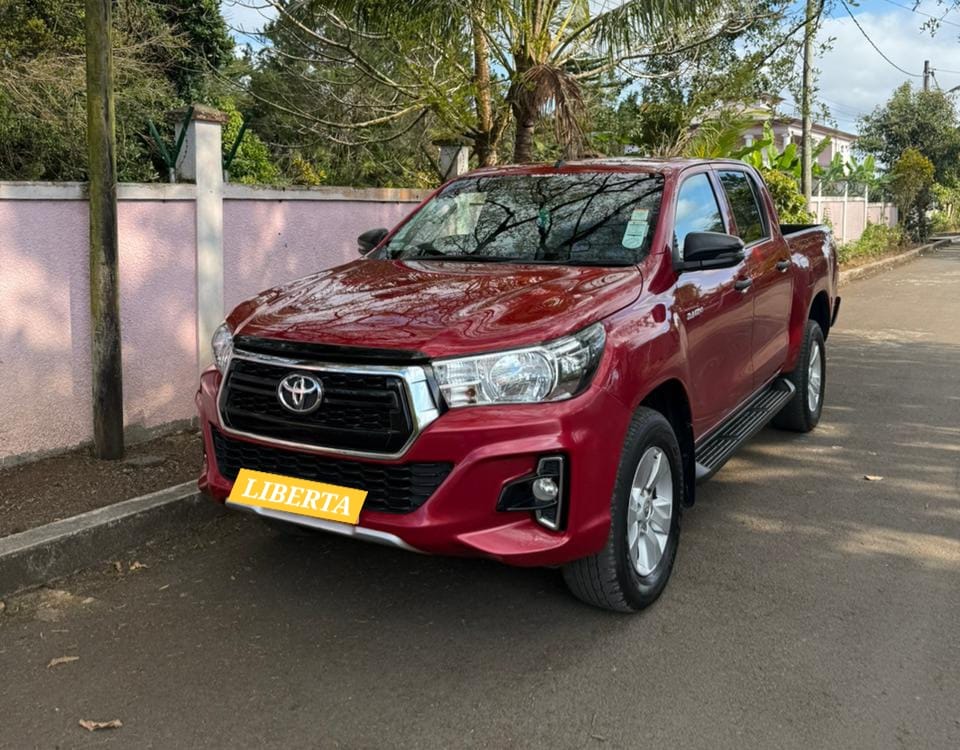 Toyota Hilux 4×4 pickup available for rent in Mauritius from Liberta Car Rental—white double-cab climbing a lush, off-road trail with mountains and palm trees in the background.