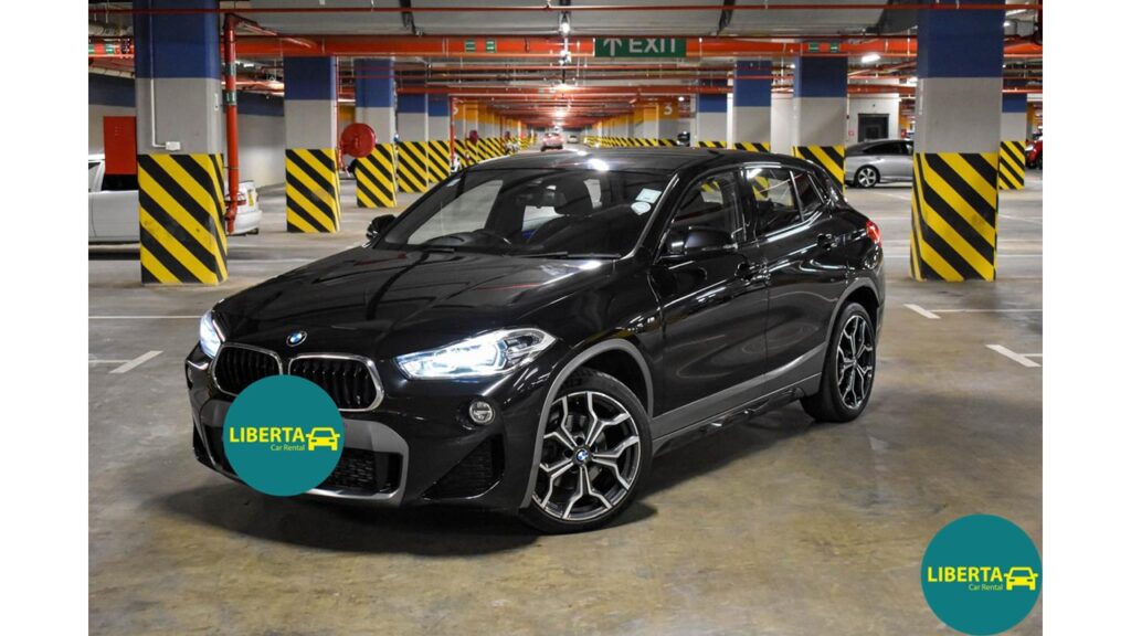 a sleek and sporty BMW X2, available for car hire in Mauritius through Liberta Car Rental. The vehicle is positioned in front of a tropical beach backdrop with palm trees and crystal-clear waters, symbolizing the perfect blend of luxury and adventure. The BMW X2’s bold grille, aerodynamic lines, and premium alloy wheels are clearly visible, showcasing its modern design. Ideal for travelers seeking a stylish and high-performance SUV to explore the island in comfort and elegance.